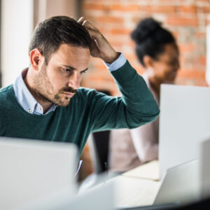 Pensive businessman reading a confusing e-mail while working on a computer in the office. His colleagues are in the background.