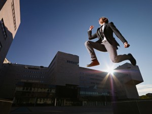 young business man jumping for joy outdoors, with office buildings and sun in background. Horizontal shape, side view, copy space