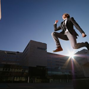 young business man jumping for joy outdoors, with office buildings and sun in background. Horizontal shape, side view, copy space